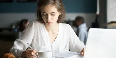 Woman writing with a drink and laptop on a desk